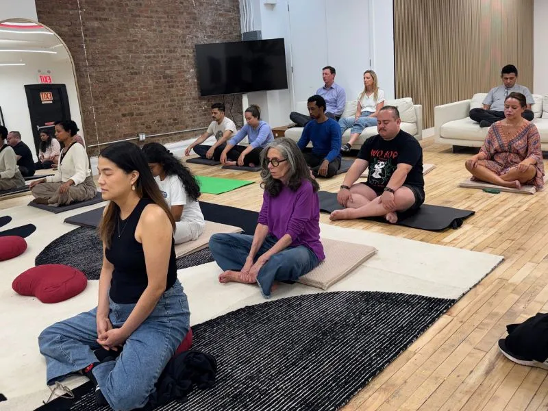 Participants meditating with eyes closed on cushions and mats in a sunlit New York loft with an exposed brick wall at the September 14 Meditate With A Monk session
