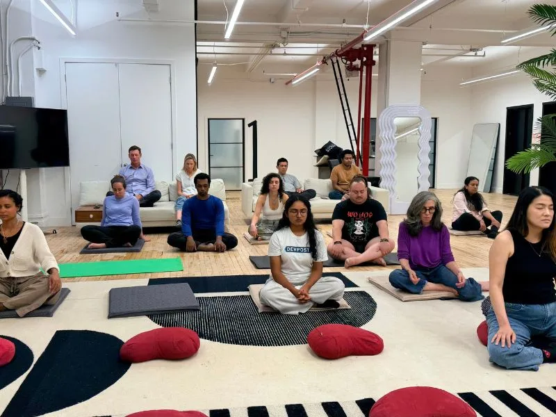 A large group meditating cross-legged on cushions and sofas in a bright white New York studio at the September 14 Meditate With A Monk session