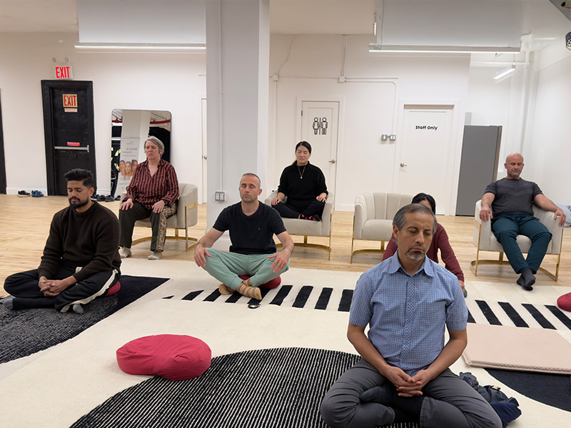 Participants meditating with eyes closed, some cross-legged on the floor and others seated in armchairs, in a bright New York studio