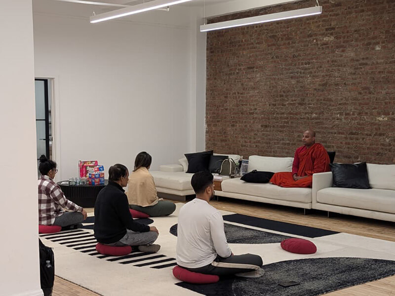 Four people seated on cushions facing a Buddhist monk in saffron robes at a Meditate With A Monk session in a Manhattan loft with an exposed brick wall