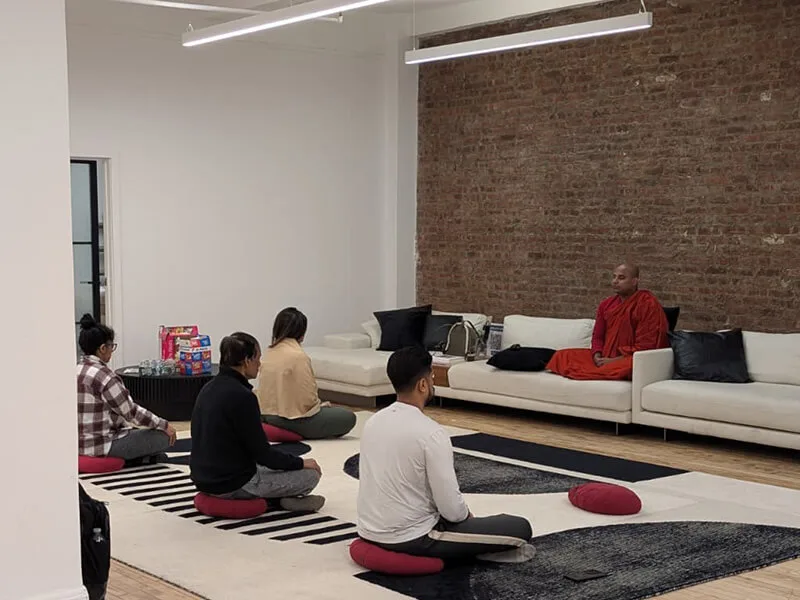 Four people seated on cushions facing a Buddhist monk in saffron robes at a Meditate With A Monk session in a Manhattan loft with an exposed brick wall